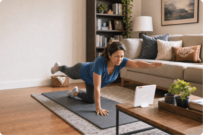 A woman exercises on a yoga mat in her living room, balancing on one knee with one arm and the opposite leg extended, while following a workout on a tablet placed on a coffee table.