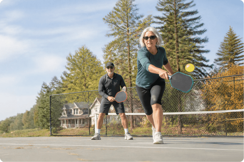 Two older adults playing pickleball outdoors on a sunny day, both holding paddles. A woman in front is reaching to hit a yellow ball, with trees and a house in the background.
