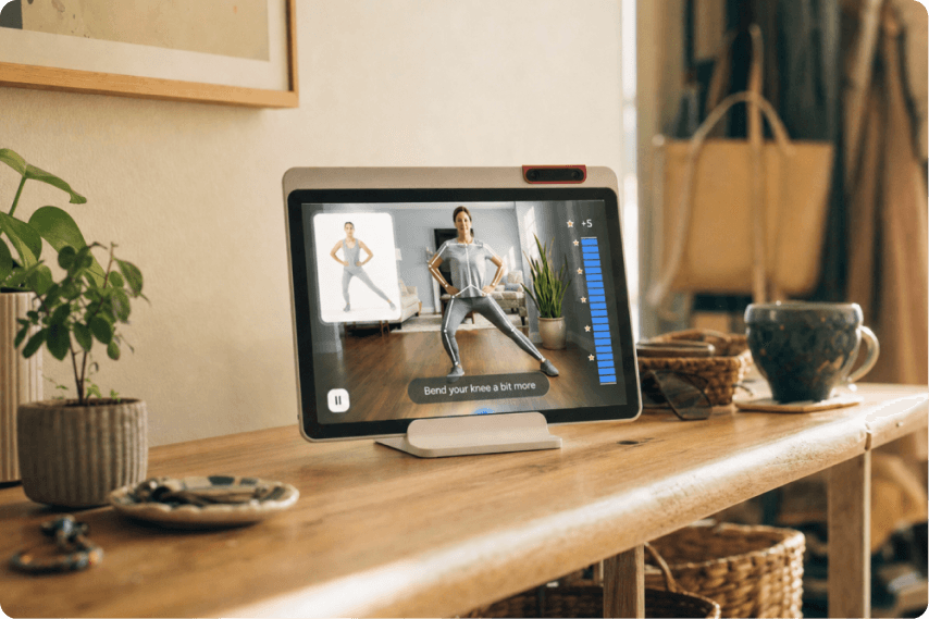 A tablet on a wooden table displays a fitness video with an instructor exercising. There are plants, baskets, and a cup nearby, creating a cozy indoor setting.