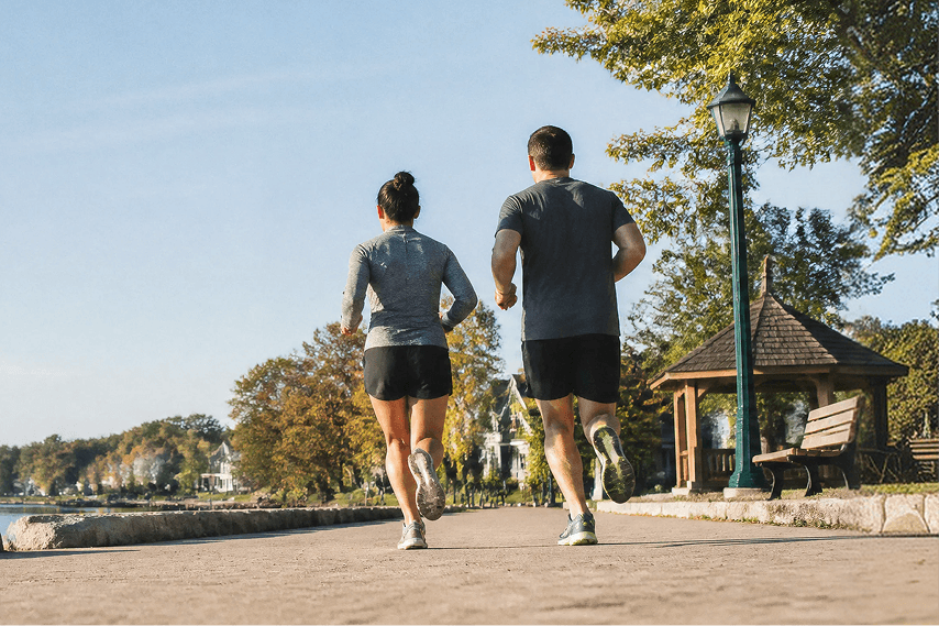 Two people in athletic clothing jog on a paved path near a park, with trees, a gazebo, benches, and houses visible in the background under a clear sky.