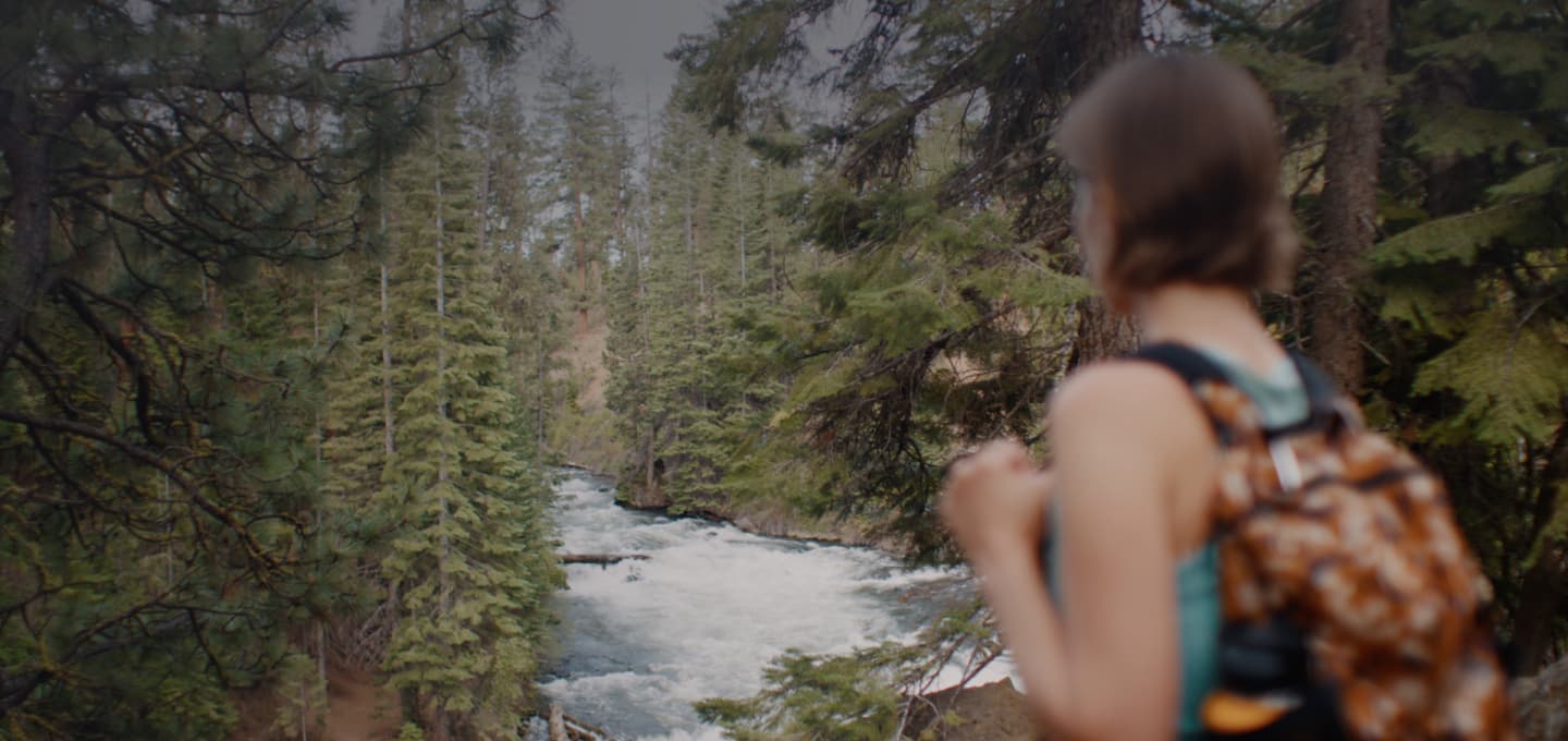 A woman with a backpack, on a hike in the woods, looking toward a river.